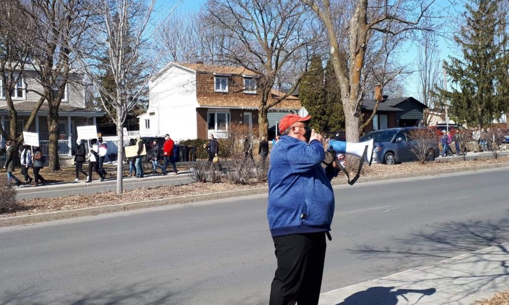 Un homme vêtu d'un manteau bleu parle dans un porte-voix alors qu'une centaine de personnes marchent derrière lui.